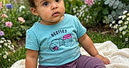 Lorraine is registered to the contest to win money with this photo: baby, child, toddler, portrait, sitting, garden, flowers, blanket, outdoor, smiling, curious, blue_shirt, purple_pants, sneakers, greenery, plants, nature, close_up, flower_bed, photography