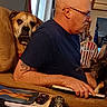 bald_man, black_dog, books, brown_dog, chair, couch, cozy, dog, dogs, furniture, glasses, indoors, living_room, man, person, pet, portrait, remote_control, sitting, television
