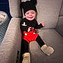 baby, child, smiling, hat, mouse_ears, red_shorts, knit_booties, pacifier, black_clothing, gray_armchair, indoor, cute, infant, seated, cozy, happy, portrait, young_child, knitwear, comfortable