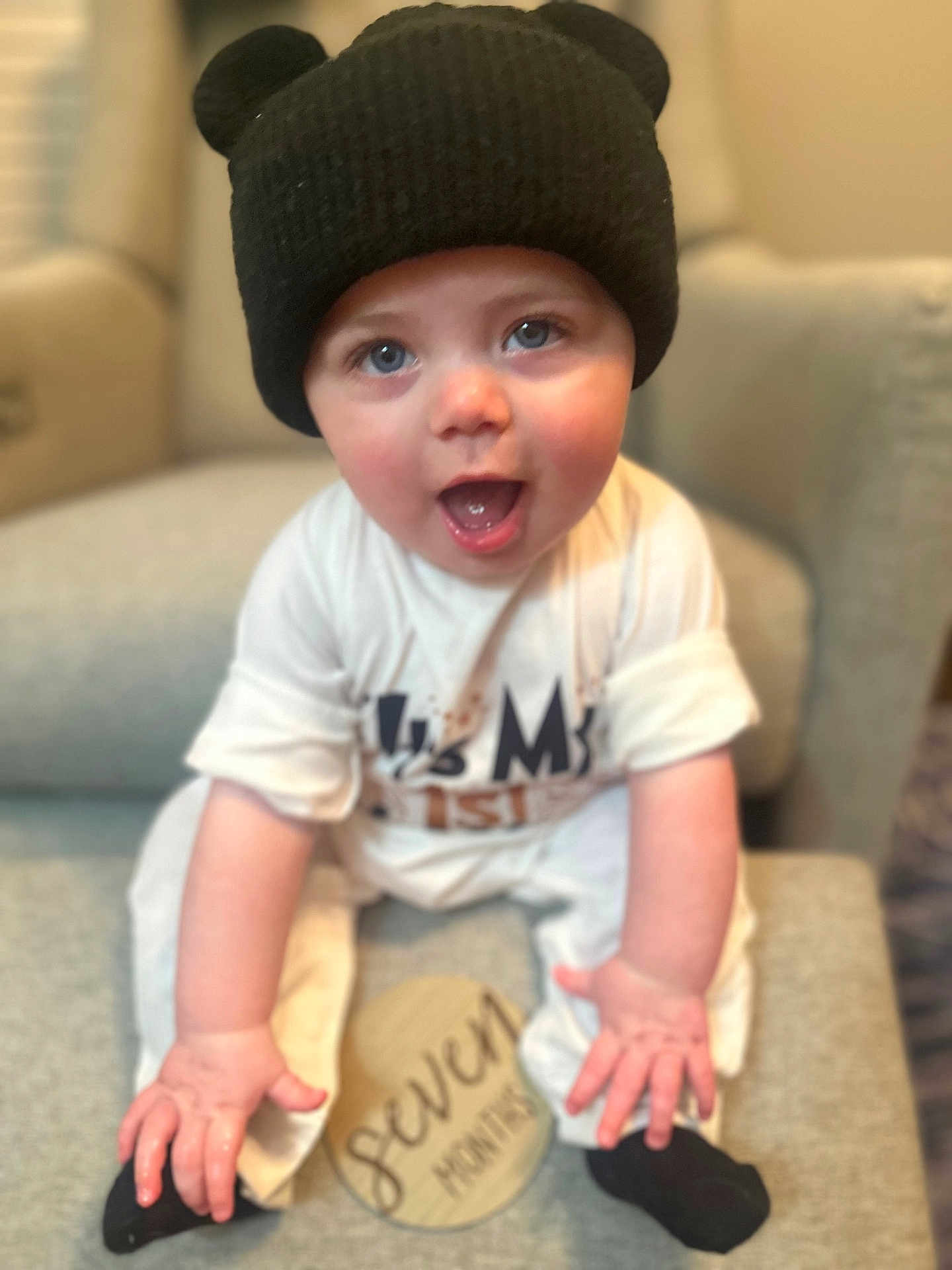 Rhett joined the competition — help win amazing prizes! baby, child, smiling, hat, black_hat, blue_eyes, cute, sitting, cushion, chair, indoor, wooden_sign, seven_months, happy, person, infant, clothing, sock, open_mouth, portrait