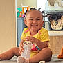toddler, smiling, child, bottle, socks, yellow_clothing, hair_buns, indoor, couch, happy, person, baby, home, furniture, high_chair, sitting, cute, playful, portrait, infant