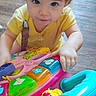toddler, child, toy, walker, yellow_shirt, blue_eyes, floor, wooden_floor, indoor, play, colorful, plastic_toy, smile, curious, young_child, person, baby, hands, closeup, activity