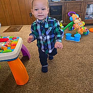 Omari is registered to the contest to win money with this photo: toddler, child, plaid_shirt, carpet, toy, indoor, cabinet, wood_paneling, colorful, walking, person, furniture, playroom, smiling, young_child, pants, socks, activity_table, plastic_toys, home