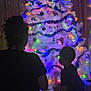 Omari is registered to the contest to win money with this photo: child, children, christmas_tree, holiday_lights, silhouette, indoor, decorations, dark, colorful, festive, tinsel, ornaments, curtains, wood_paneling, family, celebration, winter, portrait, back_view, toddler
