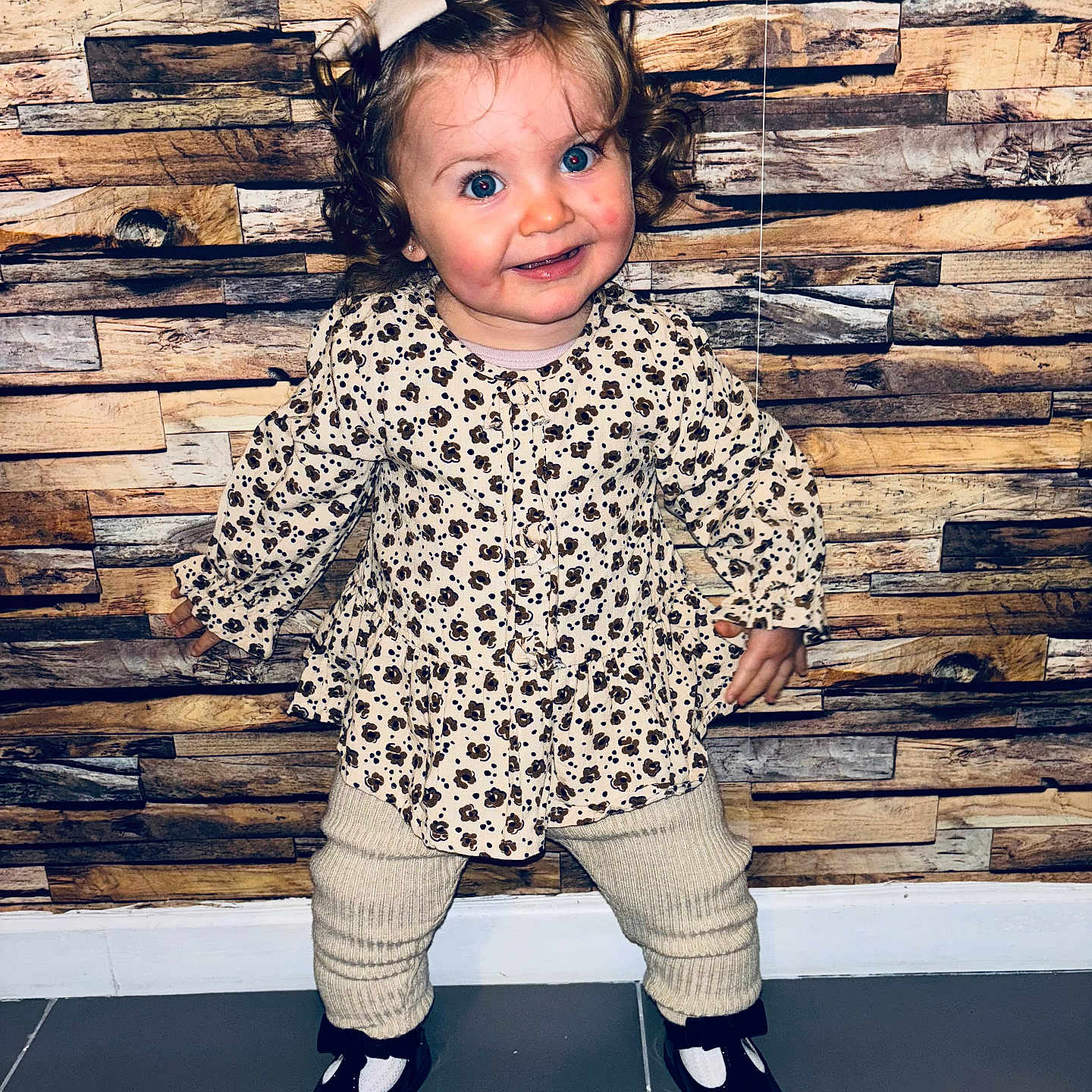 Talya participe au concours pour gagner de l'argent avec cette photo : baby, black_shoes, bow, child, clothing, curly_hair, cute, fashion, flooring, floral_top, happy, indoor, knit_pants, person, playful, portrait, smiling, standing, toddler, wooden_wall