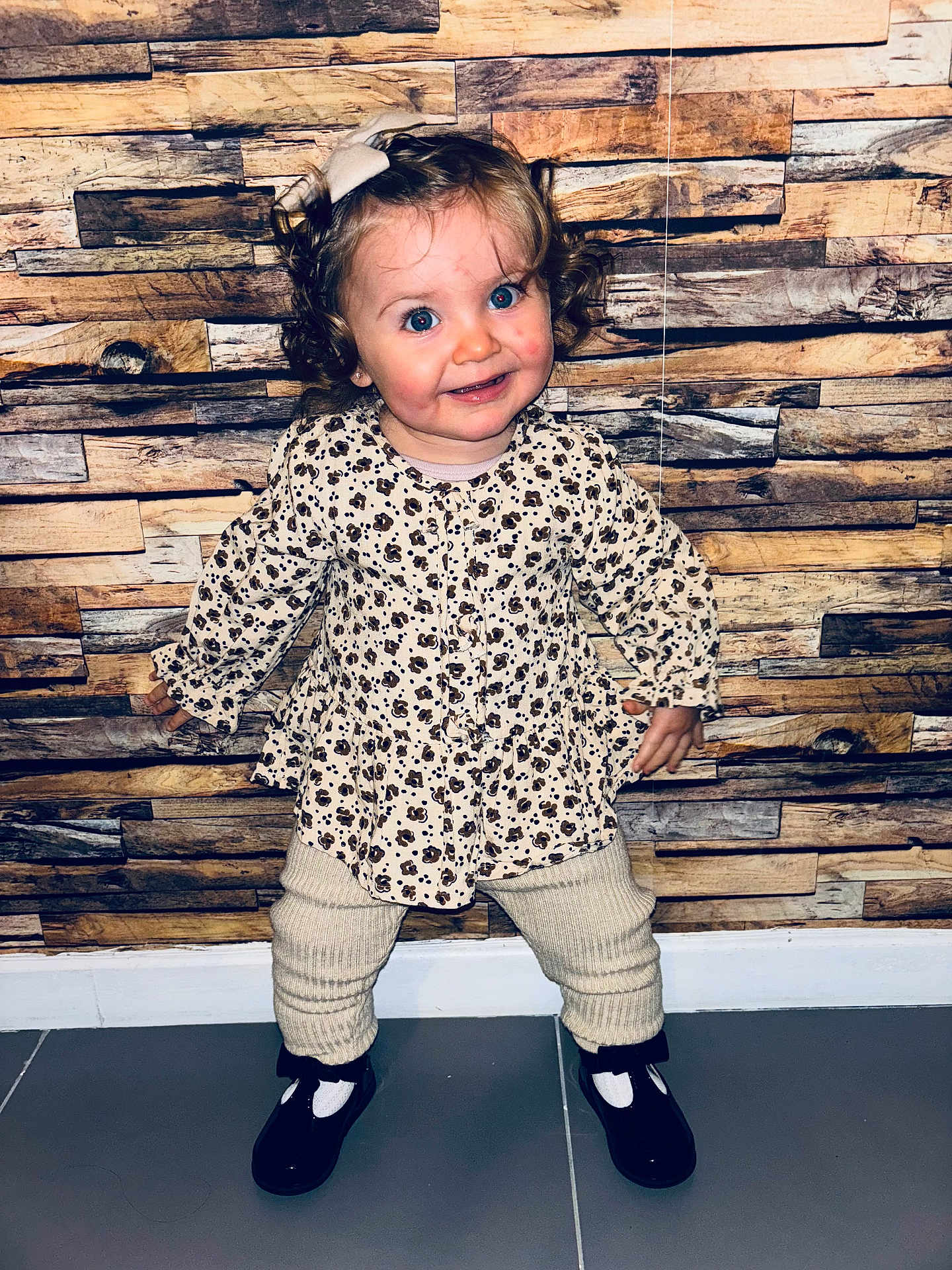Talya participe au concours pour gagner de l'argent avec cette photo : toddler, child, curly_hair, bow, floral_top, knit_pants, black_shoes, standing, smiling, indoor, wooden_wall, flooring, playful, cute, fashion, portrait, person, baby, happy, clothing