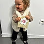 Talya participe au concours pour gagner de l'argent avec cette photo : toddler, child, baby, curly_hair, standing, indoor, white_wall, milestone_card, 9_months, vest, long_sleeve, patterned_clothing, black_pants, black_shoes, bow, smile, happy, cute, holding, floor_tiles