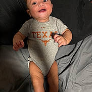 Christian is registered to the contest to win money with this photo: baby, infant, child, smiling, happy, portrait, lying_down, bed, bedding, gray_onesie, texas_logo, sportswear, nike_logo, orange_graphic, hands, legs, socks, indoor, flash_photography, close_up