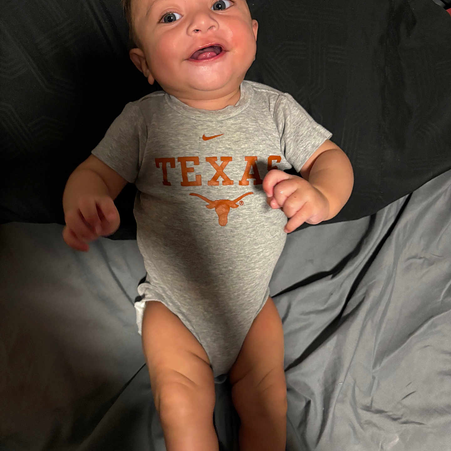 Christian is registered to the contest to win money with this photo: baby, infant, child, smiling, happy, portrait, lying_down, bed, bedding, gray_onesie, texas_logo, sportswear, nike_logo, orange_graphic, hands, legs, socks, indoor, flash_photography, close_up