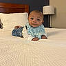 baby, infant, bed, tummy_time, bib, jeans, headboard, lamp, pillow, white_bedspread, room, indoor, child, person, cute, smiling, looking, furniture, portrait, home