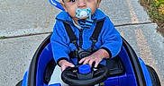 Jacob is registered to the contest to win money with this photo: baby, child, pacifier, toy_car, blue_jacket, sidewalk, grass, outdoor, sunlight, shadow, parking, car, holding, steering_wheel, cute, young_child, seatbelt, play, person, vehicle