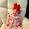 baby, child, red_bow, strawberry_pattern, cute, smiling, sitting, cream_chair, soft_texture, indoors, happy, infant, cute_outfit, headwear, plush_chair, adorable, portrait, person, young_child, cozy