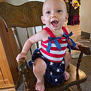 Emma joined the competition — help win amazing prizes! baby, child, smiling, happy, indoor, wooden_chair, clothing, red, white, blue, stars, stripes, kitchen, floor, carpet, foot, hand, face, person, home