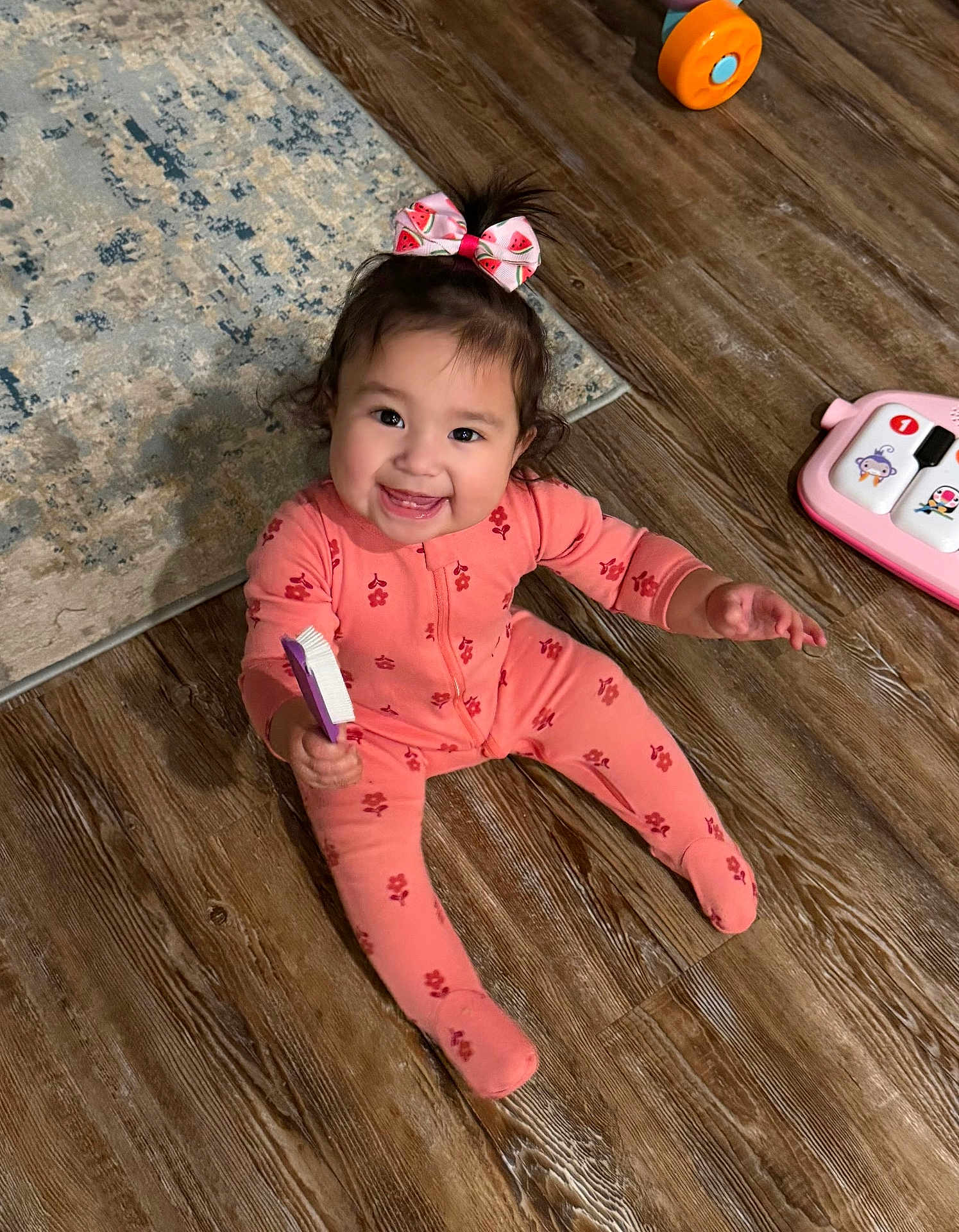Leiana is registered to the contest to win money with this photo: baby, toddler, smiling, pajamas, hair_bow, brush, toy, wooden_floor, rug, sitting, indoor, happy, face, eyes, teeth, pink, playtime, child, cute, home