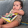 baby, child, curly_hair, soft_book, couch, gray_couch, indoor, person, infant, clothing, shorts, white_shirt, toy, colorful, seated, portrait, cute, young_child, face, hands