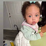 Sienna Leigh is registered to the contest to win money with this photo: baby, child, indoor, person, curly_hair, bib, light_clothing, holding, door, floor, adult, face, eyes, expression, hand, hair_bow, home, furniture, curious, portrait