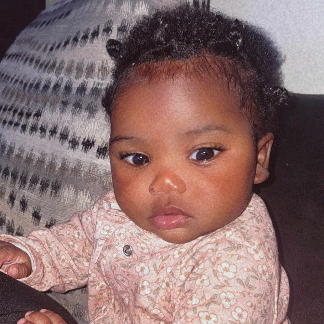 Brycelynn is registered to the contest to win money with this photo: baby, child, clothing, couch, curly_hair, cute, expression, eyes, face, floral_pattern, hair, hands, indoor, infant, person, pillow, portrait, sitting, skin, young