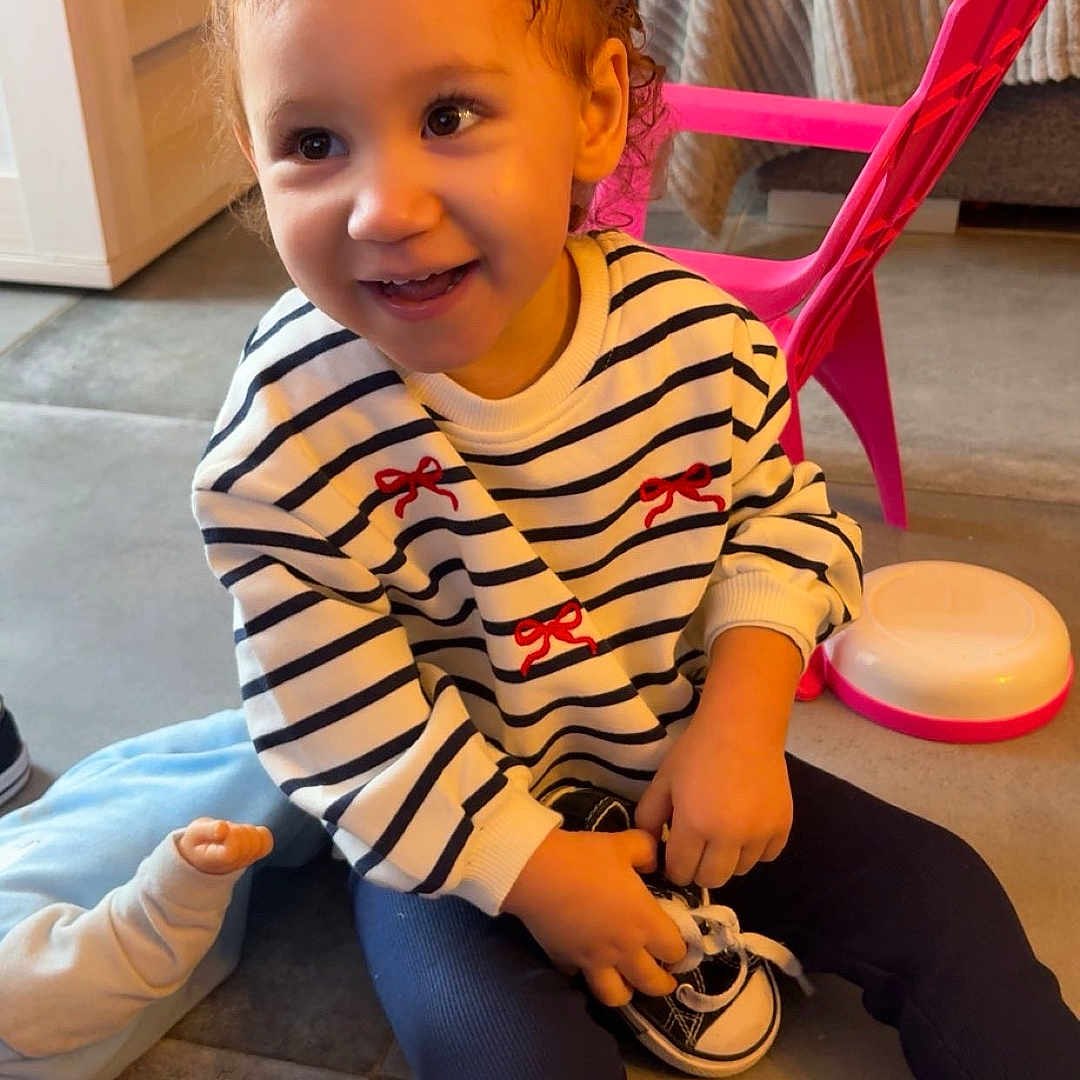 Aalyah a rejoint le concours — aidez-le/la à gagner de superbes lots ! toddler, child, smiling, striped_sweater, pigtails, indoor, floor, pink_chair, toy, couch, cushions, happy, playful, shoe, socks, person, young_child, casual_clothing, home, cute