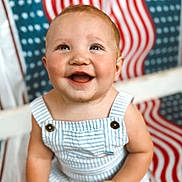 James joined the competition — help win amazing prizes! baby, child, smiling, romper, striped_clothing, sitting, happy, face, portrait, indoor, background_pattern, red, white, blue, cute, infant, person, bright, cheerful, playful