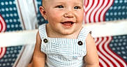 James is registered to the contest to win money with this photo: american_flag_pattern, baby, baby_skin, blond_hair, blue_eyes, chair, child, cute, face, happy, innocence, overalls, patterned_background, portrait, sitting, skin, smiling, striped_clothing, summer_clothes, young