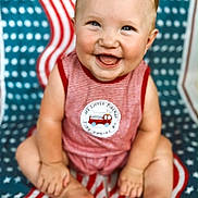 James is registered to the contest to win money with this photo: baby, child, smiling, happy, sitting, blanket, red_stripes, blue_stars, clothing, cute, portrait, infant, face, feet, hands, pattern, background, cute_baby, young_child, indoors