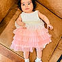 baby, child, dress, pink, white, socks, flower, couch, brown, wooden_floor, indoor, curly_hair, cute, portrait, laying_down, infant, young_child, comfortable, soft_lighting, smiling_face