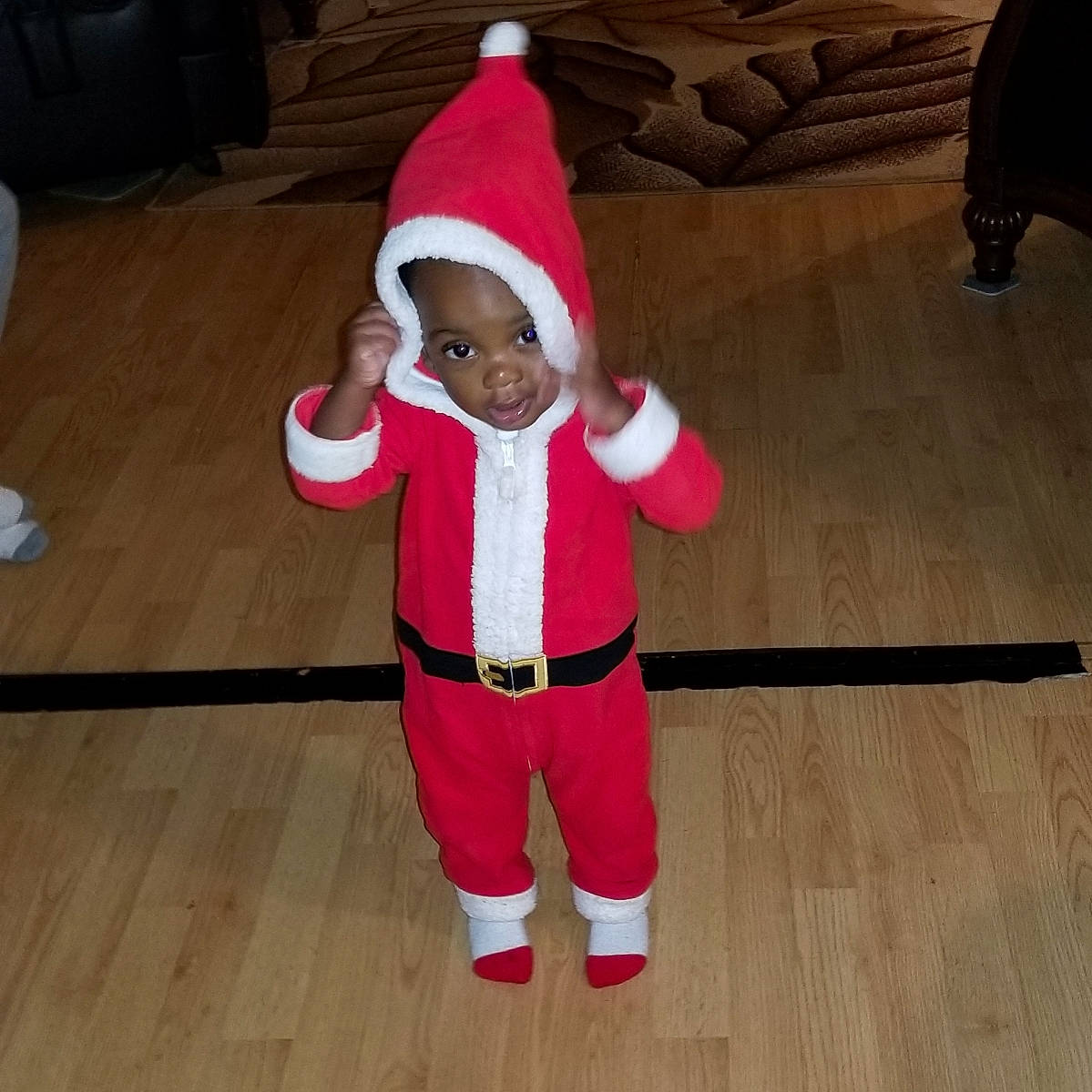 Kolby is registered to the contest to win money with this photo: baby, baby_toddler_clothing, child, christmas_decoration, costume_hat, event, fictional_character, floor, flooring, hardwood, hat, holiday, human_body, lap, person, santa_claus, sleeve, toddler, tree, wood