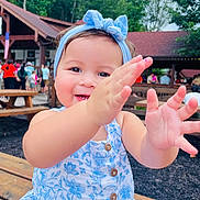 Emma is registered to the contest to win money with this photo: toddler, child, baby, smiling, blue_dress, headband, outdoor, picnic_table, people, trees, summer, playful, happy, face, hands, wooden_bench, daylight, nature, casual_clothing, reaching_out