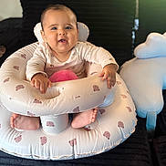 Andy is registered to the contest to win money with this photo: baby, child, infant, smiling, sitting, portrait, indoor, inflatable_seat, baby_bouncer, cushion, cloud_pillow, bed, black_blanket, pink_clothing, onesie, bare_feet, hands, face, sunlight, cozy