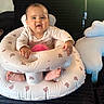 baby, child, infant, smiling, sitting, portrait, indoor, inflatable_seat, baby_bouncer, cushion, cloud_pillow, bed, black_blanket, pink_clothing, onesie, bare_feet, hands, face, sunlight, cozy