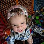 Fox is registered to the contest to win money with this photo: toddler, child, cap, smile, toy, playpen, indoor, clothing, happy, face, person, curly_hair, colorful, play_area, baby_toys, fence, cute, reaching, portrait, casual