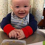 Atlus is registered to the contest to win money with this photo: baby, high_chair, bib, baby_food, spoon, smiling, happy, infant, feeding_time, cushion, striped_clothing, food_container, cute, indoors, child, person, face, hands, table, playful