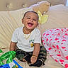 baby, smiling, sitting, bed, teddy_bear, white_tshirt, camo_pants, black_shoes, gold_jewelry, ribbons, blanket, heart_pattern, happy, child, indoor, portrait, cute, accessory, soft_toy, comfortable