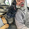 man, dog, black_dog, car, selfie, beard, smiling, open_mouth, tongue, collar, harness, seatbelt, vehicle_interior, bicycle_wheel, backseat, pet, companion, portrait, indoor, funny