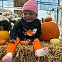 toddler, child, pumpkin, hay, orange_clothing, pink_beanie, socks, flowers, greenhouse, smiling, fall, harvest, cute, black_shirt, decor, seasonal, autumn, playful, festive, sitting