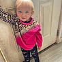 child, toddler, blonde_hair, blue_eyes, pink_sweater, leopard_print, barefoot, standing, indoors, door, floor, curious_expression, long_sleeves, pants, young_child, person, casual_pose, light, portrait, home