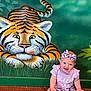 baby, child, smiling, headband, tiger_mural, wall_art, floor, colorful_clothing, sitting, indoor, cute, playful, animal_art, green_background, happy, portrait, infant, young_child, patterned_clothing, fun