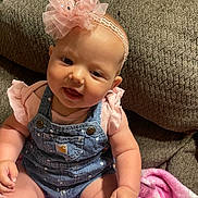 Paizley is registered to the contest to win money with this photo: baby, child, infant, denim_overalls, pink_headband, flower_accessory, couch, blanket, indoors, smiling, sitting, cute, person, skin, face, hand, furniture, clothing, headwear, happy