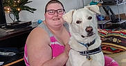 Hexiciah is registered to the contest to win money with this photo: bag, bed, blanket, christmas_tree, collar, dog, dog_tag, floor, furniture, glasses, indoor, living_room, pajamas, person, pet, shelf, sitting, smiling, string_lights, white_dog