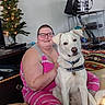 bag, bed, blanket, christmas_tree, collar, dog, dog_tag, floor, furniture, glasses, indoor, living_room, pajamas, person, pet, shelf, sitting, smiling, string_lights, white_dog