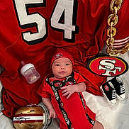 Memphis is registered to the contest to win money with this photo: baby, infant, red_clothing, football_helmet, bottle, blanket, baby_shoes, chain, sports_jersey, hat, logo, sports_fan, indoor, person, cute, laying_down, face, hand, footwear, accessory