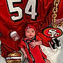 baby, infant, red_clothing, football_helmet, bottle, blanket, baby_shoes, chain, sports_jersey, hat, logo, sports_fan, indoor, person, cute, laying_down, face, hand, footwear, accessory