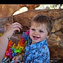 toddler, child, smiling, blue_eyes, blue_shirt, floral_pattern, beads, hand, bracelet, lei, colorful, wood_background, happy, cute, portrait, indoors, playful, baby, person, closeup