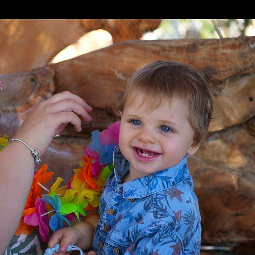 Ezio participe au concours pour gagner de l'argent avec cette photo : baby, beads, blue_eyes, blue_shirt, bracelet, child, closeup, colorful, cute, floral_pattern, hand, happy, indoors, lei, person, playful, portrait, smiling, toddler, wood_background