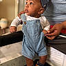 baby, child, denim_overalls, cap, sneakers, indoor, person, hand, support, curious, clothing, footwear, infant, portrait, cute, looking_away, adult_hand, soft_lighting, closeup, casual_wear