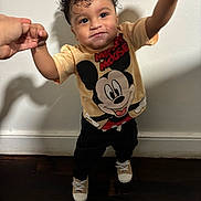Sebastian is registered to the contest to win money with this photo: toddler, child, curly_hair, mickey_mouse, tshirt, indoor, standing, holding_hands, smile, drool, person, footwear, black_pants, light_skin, adult_hands, wooden_floor, white_wall, casual_clothing, young_child, cute