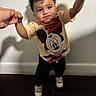 toddler, child, curly_hair, mickey_mouse, tshirt, indoor, standing, holding_hands, smile, drool, person, footwear, black_pants, light_skin, adult_hands, wooden_floor, white_wall, casual_clothing, young_child, cute