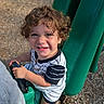 child, toddler, smiling, curly_hair, playground, swing, outdoor, portrait, happy, face, eyes, teeth, striped_shirt, shorts, shoes, seated, play_equipment, sunlight, ground, photography