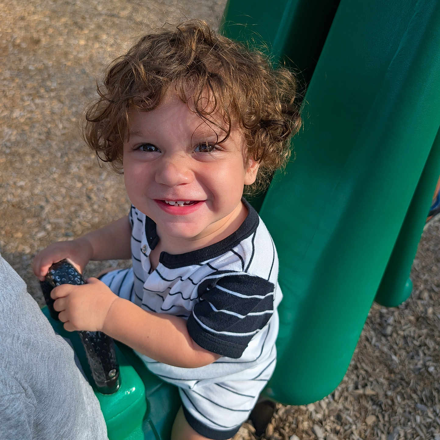 Jack joined the competition — help win amazing prizes! child, toddler, smiling, curly_hair, playground, swing, outdoor, portrait, happy, face, eyes, teeth, striped_shirt, shorts, shoes, seated, play_equipment, sunlight, ground, photography