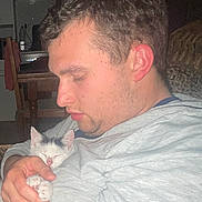 Neige a rejoint le concours — aidez-le/la à gagner de superbes lots ! man, kitten, cat, indoor, light_gray_shirt, holding, sleeping_cat, seated, gentle, cozy, face, hand, home, furniture, table, chair, peaceful, relaxed, portrait, casual